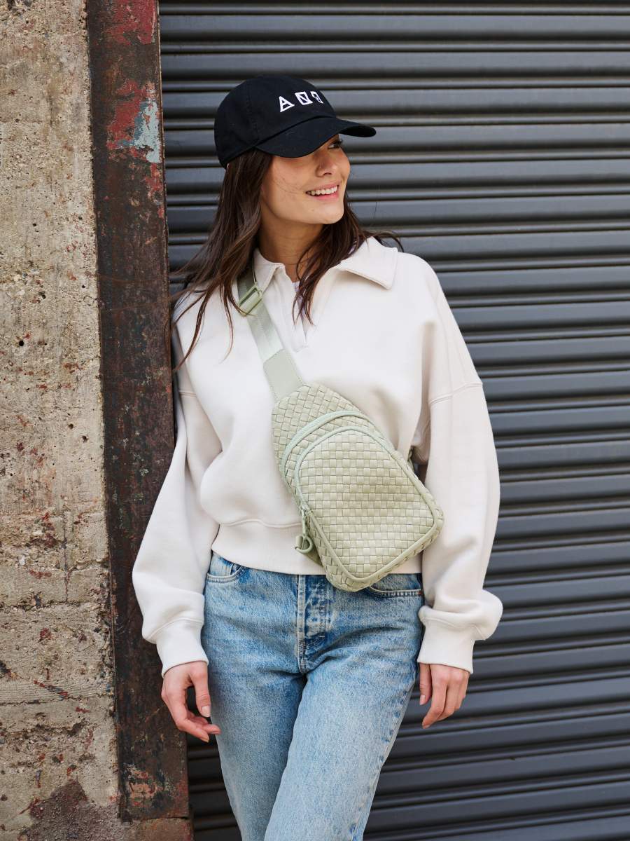 A woman wearing a cream sweatshirt, blue jeans, and a black baseball cap stands in front of a gray metal roll-up door, wearing a light green woven crossbody bag.