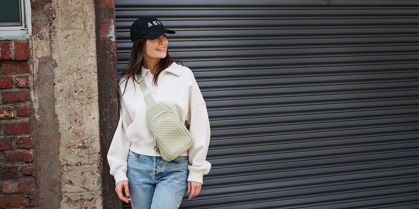 A woman wearing a cream sweatshirt, blue jeans, and a black baseball cap stands against a dark metal garage door, carrying a light green quilted crossbody bag.